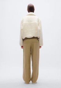 Μπεζ μπουφάν από shearling με κομψό, κομμένο σχέδιο, συνδυασμένο με χαλαρά καφέ λινά παντελόνια. Το μπουφάν διαθέτει φαρδιά γιακά και μανίκια.