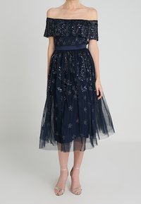 Námořnická modrá šaty bez ramínek s vyšívaným květinovým vzorem, tylová sukně a saténový pás. Vyznačuje se vrstveným, strukturovaným povrchem.