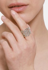 Srebrny pierścień o skomplikowanym, organicznym wzorze zwijających się gałęzi. Noszony na dłoni, prezentujący gładką, wypolerowaną teksturę.