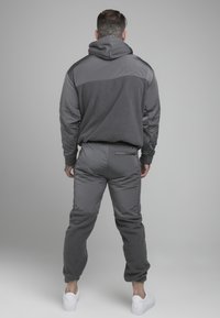 Completo sportivo grigio a due pezzi con giacca con cappuccio e zip e pantaloni con vita elasticizzata, caratterizzato da pannelli testurizzati e lisci. Sneakers bianche completano il look.