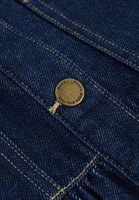 Zbliżenie na ciemnoniebieską tkaninę dżinsową z żółtym przeszyciem i mosiężnym guzikiem z napisem "Debra Dover" i "Jeans 1992."