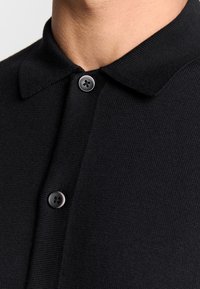 Zwarte gebreide polo met een gestructureerde kraag, vastgemaakt met twee ronde zwarte knopen. Close-up van de halslijn en knoopdetails.