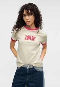 Ανοιχτό μπεζ μπλουζάκι με κόκκινη ρίγα στο λαιμό, που έχει μεγάλο κόκκινο γράμμα "LOVE" και σχέδιο τριαντάφυλλου. Φοριέται με σκουρό μπλε τζιν.