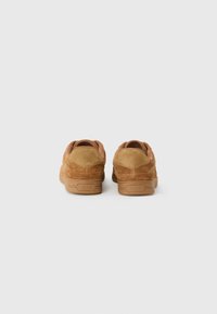 Tan suede sneakers with a smooth texture and rubber soles, featuring a rounded back and embossed brand logo on the heel.
