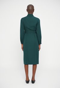 Roheline vöötohvel dress, millel on pikad varrukad, krae ja sirge lõige. Sile tekstuur ja põlvekõrgune äär. Mustad lamedad kingad täiendavad komplekti.