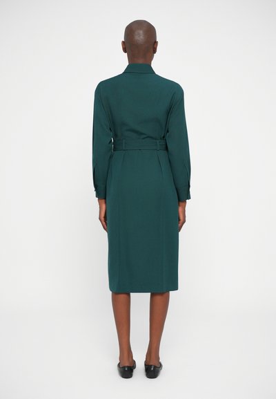 Robe verte ceinturée avec des manches longues, un col et une coupe droite. Texture lisse et longueur au genou. Des chaussures plates noires complètent la tenue.