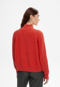 Gebreide rode trui met een hoge kraag, geribbelde textuur en verlaagde schouders, gecombineerd met aansluitende grijze broek.