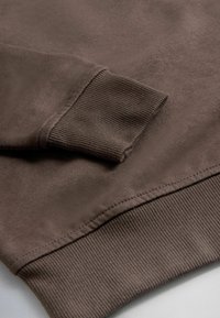 Barna pulóver anyagának közeli képe, amelyen látható a bordázott mandzsetta és az alsó szegély varrási részletei.