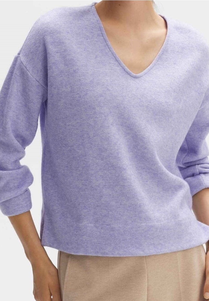 Personne portant un pull à col en V violet clair avec les manches retroussées et un pantalon beige, mains partiellement dans les poches.