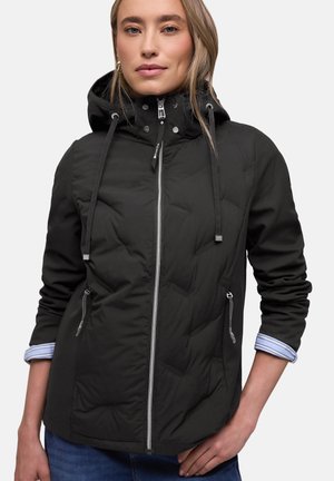 Femme portant une veste à capuche noire avec fermeture éclair à l'avant et poches zippées, manches retroussées pour révéler les poignets de chemise à rayures.