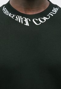 Pogled od blizu na crnu majicu s okruglim izrezom na kojoj je bijeli natpis "VERSACE JEANS COUTURE" ispisan oko izreza na gornjem dijelu prsa osobe.