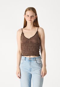 Brown cropped top with ruffled edges and smocked texture, featuring thin straps and a V-neck design. Paired with light blue jeans.