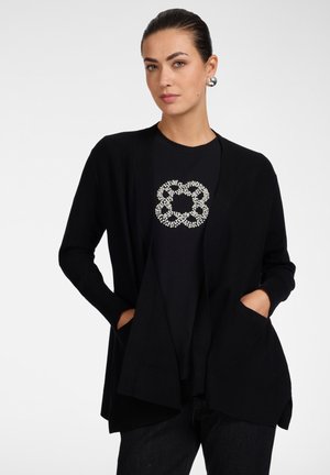 Frau trägt eine schwarze Strickjacke über einem schwarzen Hemd mit weißem Perlenblumenmuster, Hände in den Taschen und silberne runde Ohrringe.