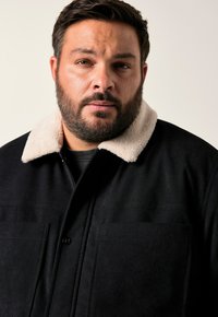 Svart jacka med en beige, mjuk pälskrage, som har två framfickor och knäppning med knappar, tillverkad av texturerat tyg.