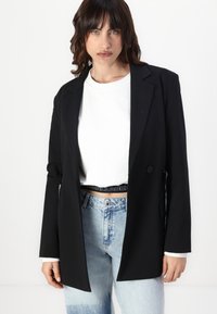 Zwarte blazer met een nauwsluitende pasvorm, twee knopen en een zachte textuur. Draagt over een witte shirt met een grafische zwarte tailleband en lichtblauwe spijkerbroek.