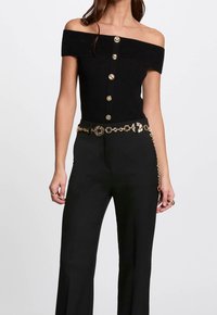 Černý top bez ramínek s žebrovaným vzorem a zlatými knoflíky, v kombinaci s černými kalhotami s širokými nohavicemi a zlatým opaskem s řetězem, který má kruhové články.