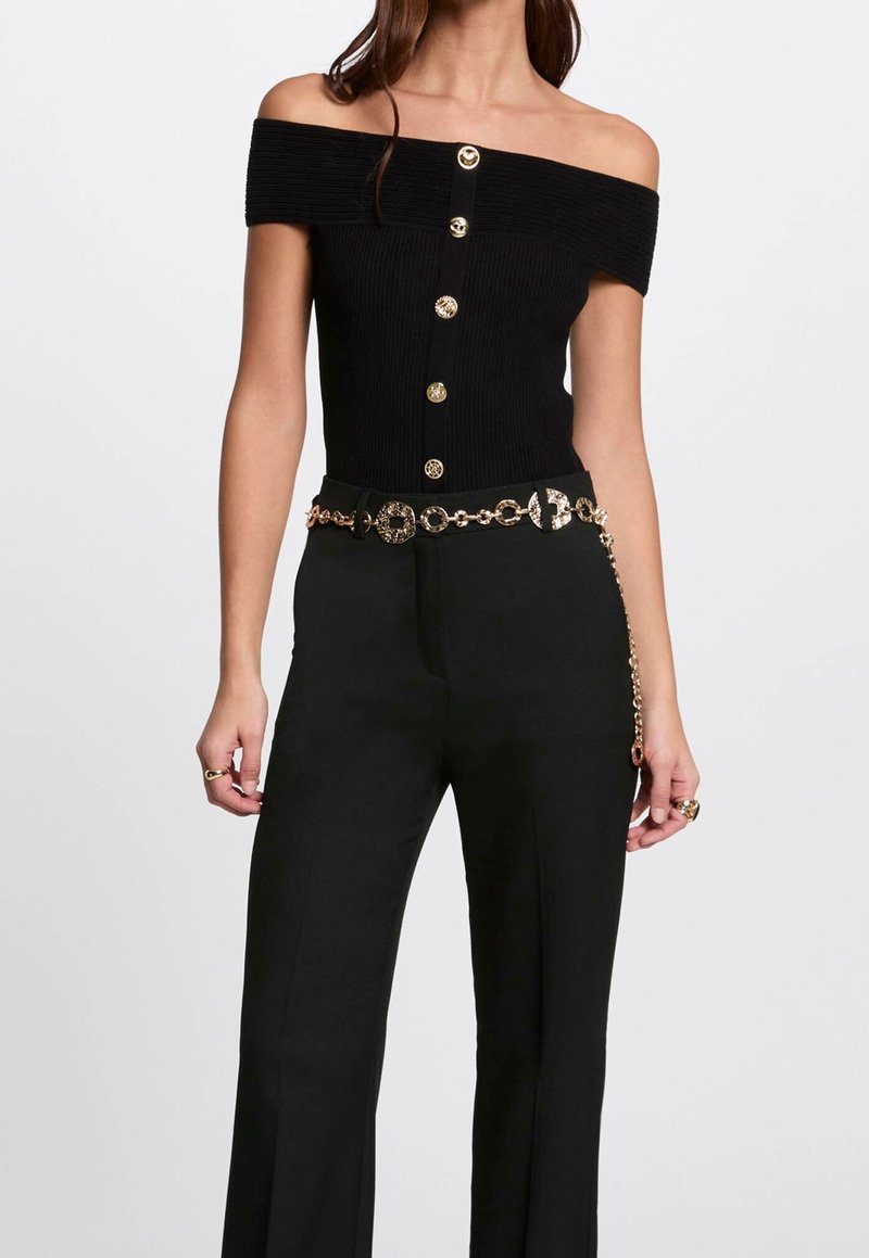 Černý top bez ramínek s žebrovaným vzorem a zlatými knoflíky, v kombinaci s černými kalhotami s širokými nohavicemi a zlatým opaskem s řetězem, který má kruhové články.