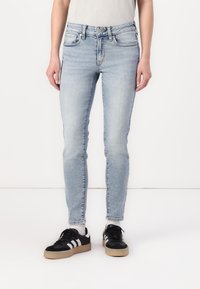 Jeans skinny de color azul claro con una cintura alta, diseño clásico de cinco bolsillos y un ligero desgaste. Combinados con zapatillas negras con rayas blancas.