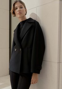 Giacca nera doppiopetto in tessuto strutturato con ampi rever, tasche laterali e dettagli con bottoni. Indossata sopra un maglione nero a maglia.