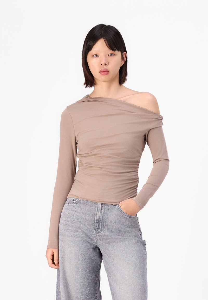 Top cu mâneci lungi, off-the-shoulder, dintr-un material bej moale, cu detalii pliate; asortat cu blugi gri cu talie înaltă.
