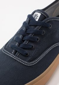Sneaker in tela navy con cuciture bianche a contrasto, occhielli neri, lacci rotondi e suola in gomma marrone chiaro. Logo circolare sulla linguetta.