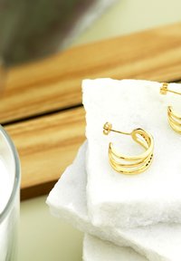 Orecchini a cerchio in oro con tre bande curve, caratterizzati da superfici lucide. Presentati su pietre di marmo bianco vicino a una candela di vetro.