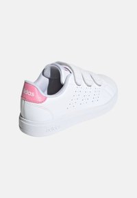 Zapatilla blanca con parte superior de cuero suave, detalles perforados y un acento rosa en el talón. Presenta una suela de goma plana y correas de Velcro.