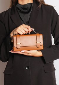 Borsa in ecopelle marrone con accenti dorati, forma rettangolare, manico superiore e dettagli con doppia fibbia. Tenuta in mano contro un outfit nero.