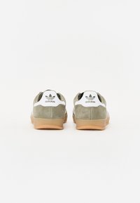 Adidași din suede verde cu un tab heel alb, talpă din cauciuc de culoare guma și o dâră neagră. Caracteristici include logo-ul Adidas pe călcâi și pe lateral.