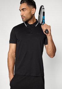 Černé polo tričko s bílými pruhy na límci, krátkými rukávy a logem na hrudi. Model drží tenisovou raketu s modrým úchopem v druhé ruce.