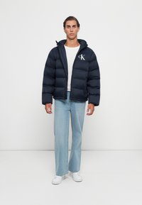 Man in een donkere pufferjack met "CK" logo, wit T-shirt, lichtblauwe wijde jeans en witte sneakers die tegen een witte achtergrond staat.