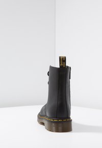 Bota de tobillo de cuero negro con costuras amarillas, suela translúcida con estrías y un tirador con texto amarillo, mostrada desde la parte posterior sobre una superficie blanca.