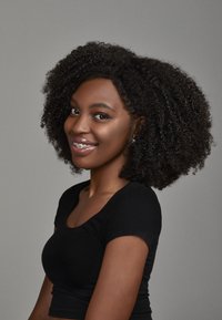 Portrait einer Person mit voluminösem, lockigem Haar und Zahnspangen, die ein enganliegendes, schwarzes kurzärmeliges Shirt vor einem neutralen grauen Hintergrund trägt.