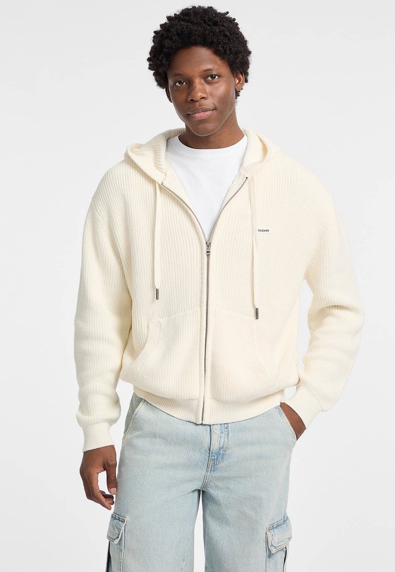 Kremasti zip-up dukserica s rebrastom teksturom, oversize kroj, prednje džepove i podesive vezice. Nositi preko bijele majice.