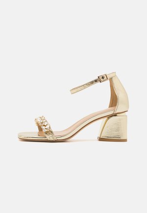 Sandalias de tacón metálicas doradas con una tira ancha adornada con piedras de strass transparentes, puntera cuadrada y tacón grueso. Correa ajustable en el tobillo.