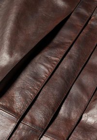 Bruin lederen stof met een gladde textuur, gekenmerkt door geplooide plooien en subtiele variaties in kleur en glans. Minimale stikdetails zichtbaar.