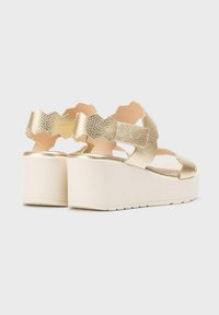 Sandalias de plataforma de imitación cuero dorado con un diseño de tira ondulada, suelas blancas contrastantes y acentos texturizados.