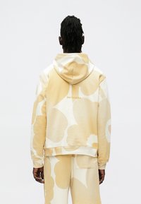 Sweatshirt com capuz em tecido bege suave com grandes padrões florais em creme claro. Logo impresso nas costas. Corte descontraído com punhos canelados.