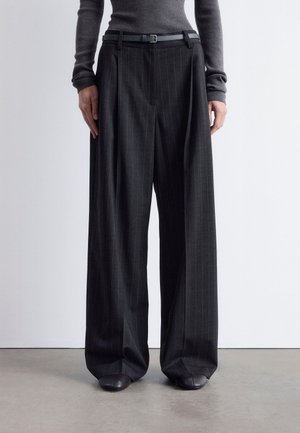 Person wearing high-waisted, wide-leg black pinstripe trousers with a black belt and dark shoes, standing against a plain white background.