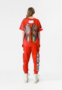 Camiseta roja con diseño de alas coloridas en la parte posterior, pantalones deportivos rojos a juego con acentos blancos y botas color crema. Etiqueta adjunta a los pantalones.