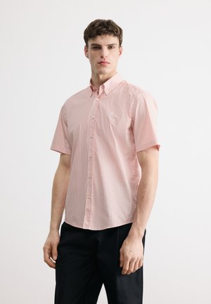 Ung mand iført en kortærmet, pink og hvidstribet skjorte med knapper og sorte bukser, stående mod en ensfarvet hvid baggrund.