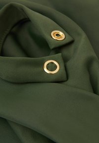 Gros plan sur un tissu vert foncé montrant deux œillets en métal doré sur des couches pliées de matériau texturé.