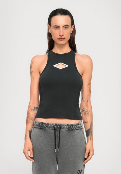 Žena s dugom smeškom kestenjastom kosom nosi crni bezrukavni crop top s rezom u obliku dijamanta i sivi dukserica s vezicama, stojeći na bijeloj pozadini.