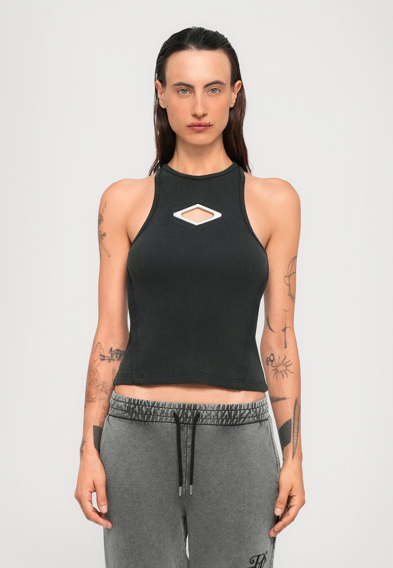 Woman with long brown hair wears black sleeveless crop top with diamond-shaped cutout and gray drawstring sweatpants, standing against white background.
