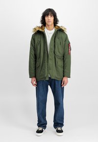Mann med krøllete hår som har på seg en olivengrønn parkas med pelshette, hvit skjorte, blå jeans, og svart-hvite sneakers som står mot en hvit bakgrunn.