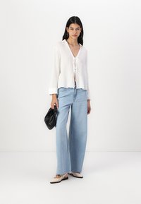Bluză albă, texturată, cu un detaliu legat în față, completată cu blugi din denim ușori, cu talie înaltă și pantaloni largi de culoare albastru deschis. O poșetă neagră și pantofi cu două tonuri finalizează ținuta.