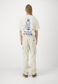 Tricou din bumbac bej cu un grafic imprimat cu o pisică albastră pe spate. Combinate cu pantaloni cargo bej. Ambele au croieli relaxate.