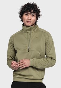 Olivgrüner Pullover mit hohem Kragen, Frontreißverschluss und einer Brusttasche. Weicher Stoff mit einem Logo auf der oberen linken Seite und an den Ärmeln. Schwarze Hose.