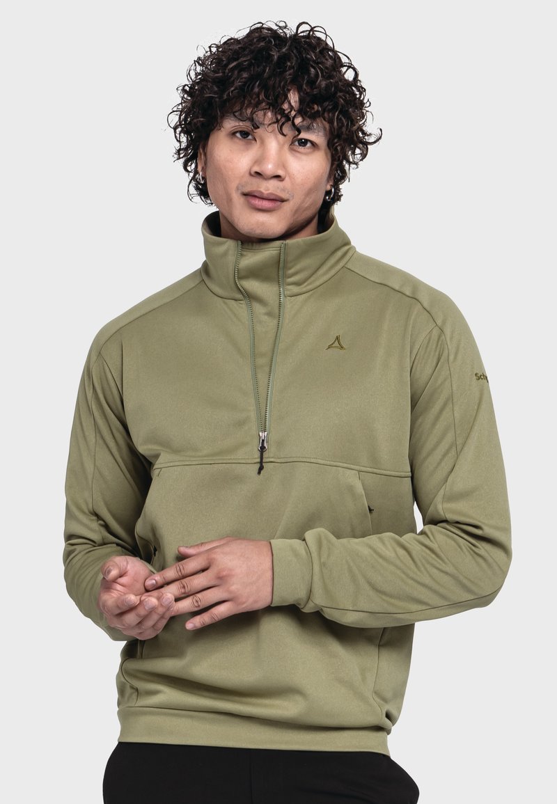 Olivgrüner Pullover mit hohem Kragen, Frontreißverschluss und einer Brusttasche. Weicher Stoff mit einem Logo auf der oberen linken Seite und an den Ärmeln. Schwarze Hose.