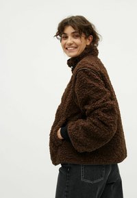 Giovane donna sorridente, che indossa una giacca di pile marrone a trama con le mani nelle tasche, abbinata a jeans scuri, in piedi davanti a uno sfondo semplice.